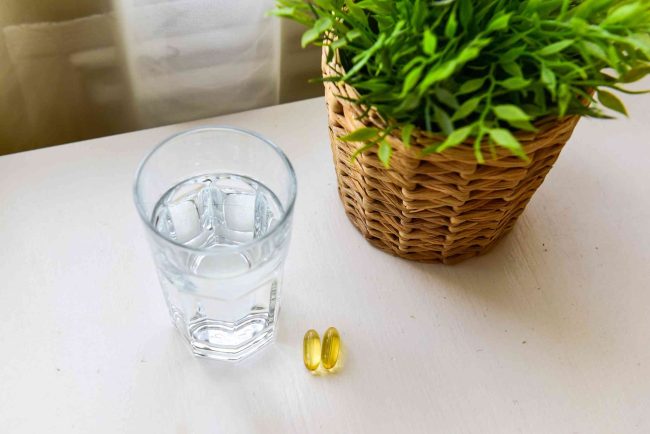 Glass of water and two chlorophyll capsules laid on a white table with a woven pot of a green plant.